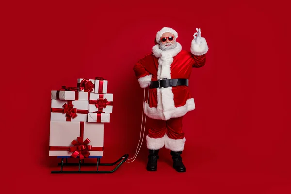 Full length photo of funky santa show okey bring gifts by sled wear cap glasses coat pants shoes απομονωμένα σε φόντο κόκκινο χρώμα — Φωτογραφία Αρχείου