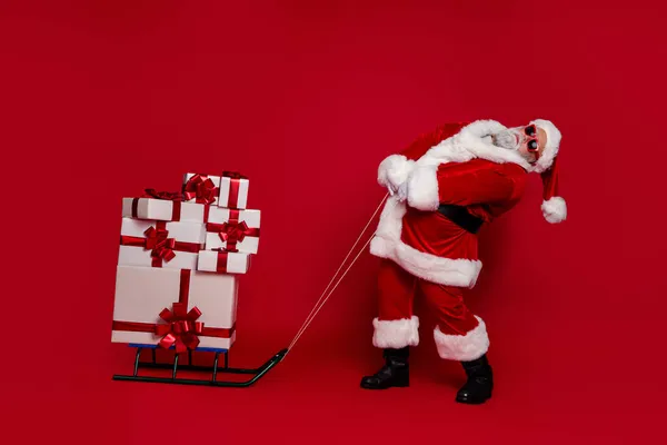 Fotografie silného funky Santa Claus nosit červený kostým klobouk tmavé brýle tahání sáně dárky zásobník izolované červené barvy pozadí — Stock fotografie