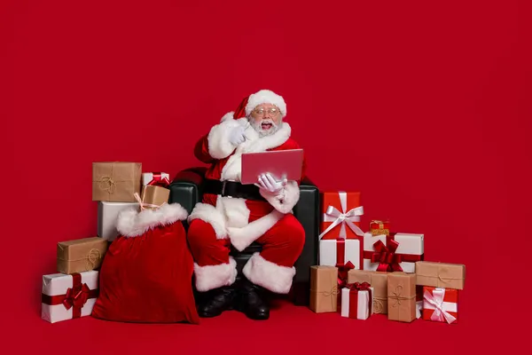 Full size foto van onder de indruk santa sit geschenken kopen online index laptop dragen pet bril broek schoenen geïsoleerd op rode achtergrond — Stockfoto