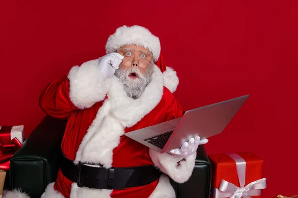 Foto de impressionado santa sit look laptop desgaste chapéu casaco de óculos isolado no fundo vermelho — Fotografia de Stock