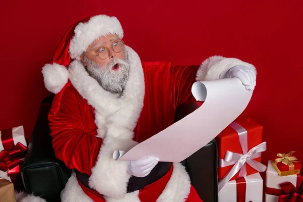 Foto de Papai Noel ler carta desejo perto apresenta desgaste chapéu casaco de óculos isolado no fundo vermelho — Fotografia de Stock