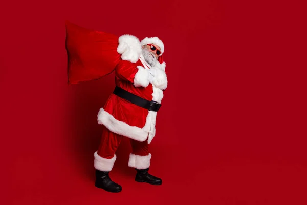 Foto de perfil de longitud completa de sobrepeso santa llevar bolso grande con regalos desgaste sombrero gafas abrigo pantalones botas aisladas sobre fondo rojo —  Fotos de Stock