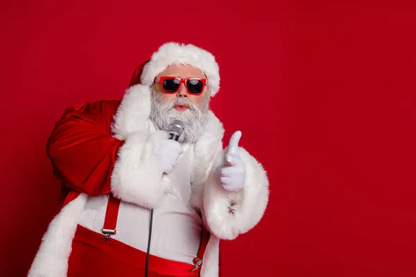 Foto di cool divertente Babbo Natale indossare costume rosso occhiali scuri canto karaoke indicando dito vuoto spazio isolato colore rosso sfondo — Foto Stock