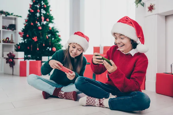 Zdjęcie dwóch graczy dzieci trzymać telefon cieszyć X-mas gry wideo wydarzenie nosić jumper kapelusz w urządzonym domu w pomieszczeniach — Zdjęcie stockowe
