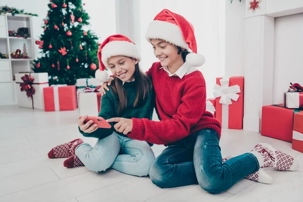 Zdjęcie dwojga słodkich dzieci przytulić trzymać telefon zegarek x-mas film nosić sweter kapelusz w urządzone domu w pomieszczeniach — Zdjęcie stockowe