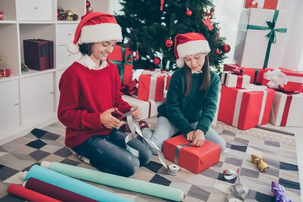 Foto de niños positivos ayudan a santa sentarse piso preparar diciembre presenta desgaste x-mas sombrero jersey en casa decorada en interiores —  Fotos de Stock
