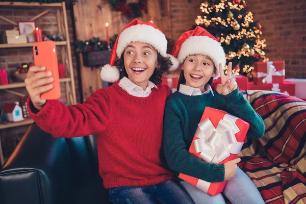 Porträtt av attraktiva glada syskon sitter på divan innehav presentförpackning med selfie visar v-tecken på dekorerad inredning hem inomhus — Stockfoto