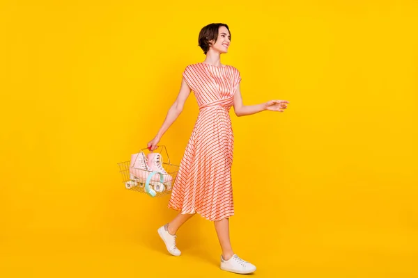 Profiel foto van positieve vreedzame meisje lopen dragen mand slijtage gestreepte jurk schoenen geïsoleerde gele kleur achtergrond — Stockfoto