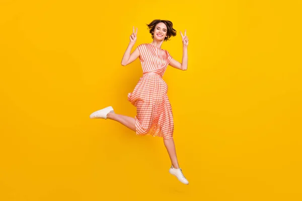 Foto von positiven freundlichen Mädchen Sprung zeigen zwei V-Zeichen tragen gestreifte Kleid Turnschuhe isoliert gelbe Farbe Hintergrund — Stockfoto