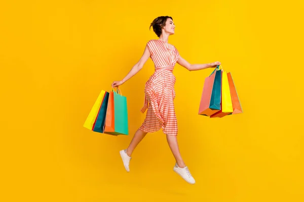 Foto de perfil de despreocupado menina salto transportar loja sacos desgaste listrado vestido tênis isolado cor amarela fundo — Fotografia de Stock