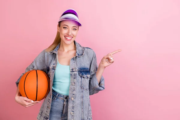 Foto de alegre millennial senhora ativa segurar ponto bola espaço vazio desgaste calça jeans camisa cap isolado no fundo cor-de-rosa — Fotografia de Stock