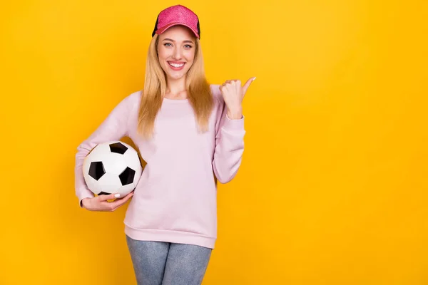 Photo of pretty adorable kobieta ubrana różowy pullover głowy trzymając piłkę wskazując kciuk puste miejsce izolowane żółty kolor tło — Zdjęcie stockowe