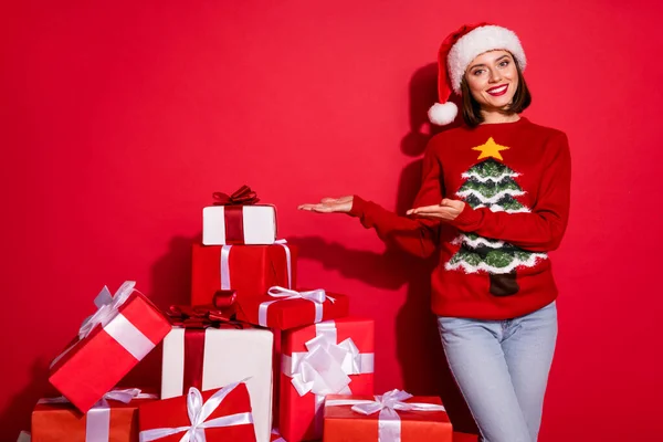 Фото чарівної блискучої молодої леді, одягненої в друк пуловер готує різдвяні подарунки, тримаючи руки порожнім простором ізольований червоний колір фону — стокове фото