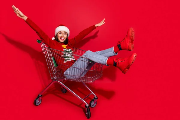 Foto lateral de perfil de cuerpo completo de la joven alegre niña sentada carrito de la tienda espíritu de adviento aislado sobre fondo de color rojo — Foto de Stock