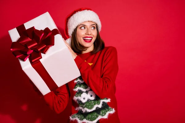 Φωτογραφία από αρκετά εντυπωσιασμένη νεαρή κοπέλα ντυμένη εκτύπωση πουλόβερ κρατώντας δώρο Χριστουγέννων αναζητούν κενό χώρο απομονωμένο φόντο κόκκινο χρώμα — Φωτογραφία Αρχείου