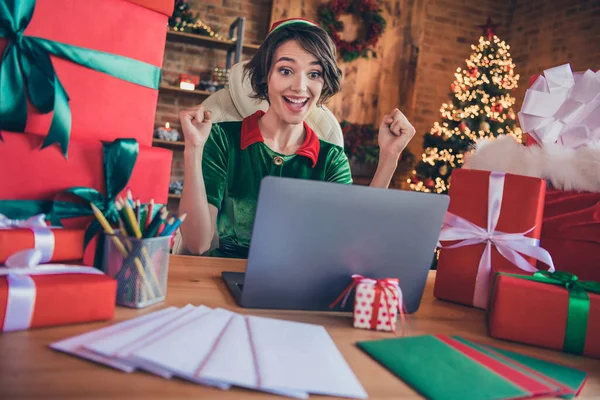 Fénykép a fiatal, nagyon vidám boldog Mikulás segítő manó ül íróasztal karácsony megjelenés laptop xmas nyerni beltéri házban otthon — Stock Fotó