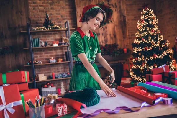 Фото чарівної красивої молодої жінки, одягненої в зелений костюм посміхаючись упаковки різдвяного подарункового різання паперу в приміщенні будинку — стокове фото