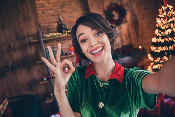 Foto di piuttosto divertente giovane donna vestita di verde costume sorridente che celebra il nuovo anno mostrando okey casa in casa stanza — Foto Stock