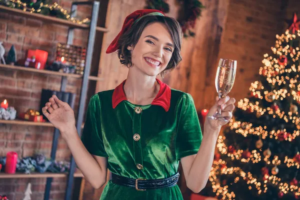 Фото блестящей милой молодой женщины, одетой в зеленый костюм, улыбающейся, празднуя Новый год, пьющей шампанское в помещении дома — стоковое фото