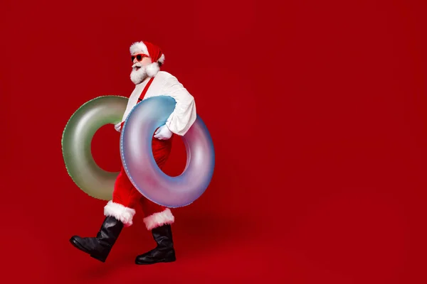 Heyecanlı, komik, emekli, Noel Baba kostümü giymiş, elinde cankurtaran şamandıraları olan, boş alanda yürüyen, izole edilmiş kırmızı arka plan fotoğrafçısı. — Stok fotoğraf