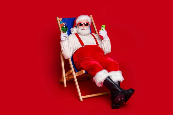 Foto de homem aposentado muito engraçado usar o traje de Papai Noel sentado cadeira de praia desfrutando de coquetel digitando moderno dispositivo isolado cor vermelha fundo — Fotografia de Stock