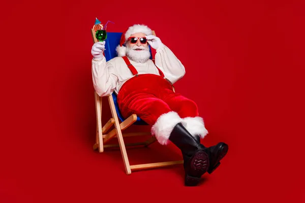 Фото крутого фанк-чоловіка пенсіонера одягненого в костюм Санти Клауса, який сидить на стільці, п'є коктейль, посміхаючись ізольований червоний колір фону — стокове фото