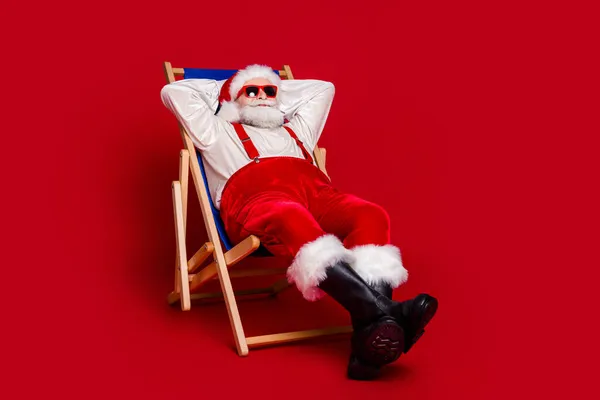 Foto van vrolijke mooie gepensioneerde man dragen kerstman kostuum glimlachen zittende ligstoel armen achter hoofd geïsoleerde rode kleur achtergrond — Stockfoto