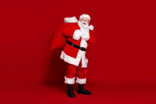 Foto de homem animado engraçado pensionista vestido traje de Papai Noel carregando grande saco de Natal sorrindo isolado fundo de cor vermelha — Fotografia de Stock