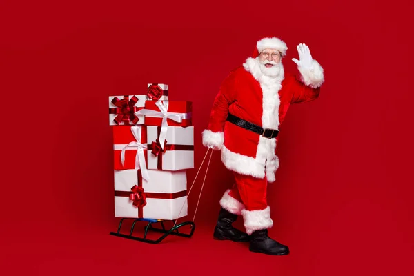 Foto de homem aposentado alegre funky usar óculos de traje de Papai Noel arrastando braço acenando Natal apresenta isolado fundo cor vermelha — Fotografia de Stock