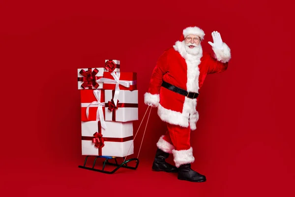 Foto de funky alegre jubilado hombre desgaste santa claus traje gafas arrastrando agitando brazo navidad presenta aislado color rojo fondo —  Fotos de Stock