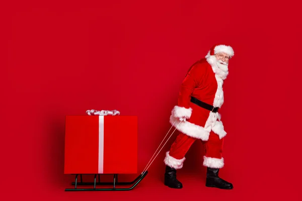 Φωτογραφία του funky κουρασμένος συνταξιούχος ντυμένος Santa Claus κοστούμι σέρνοντας μεγάλο χριστουγεννιάτικο δώρο χαμογελώντας απομονωμένο φόντο κόκκινο χρώμα — Φωτογραφία Αρχείου