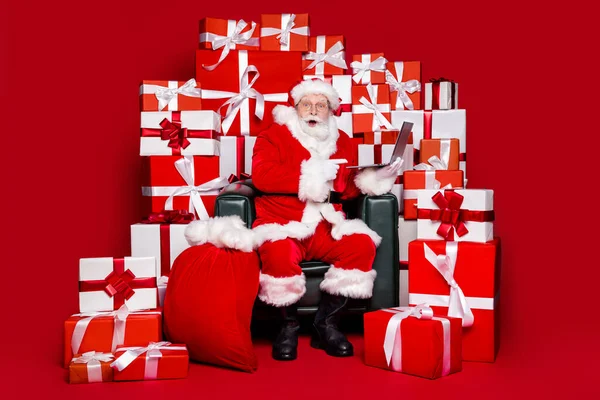 Фото веселого впечатлённого пенсионера в костюме Санта Клауса, готовящего новогодние подарки, указывающие на изолированный красный цвет ноутбука — стоковое фото