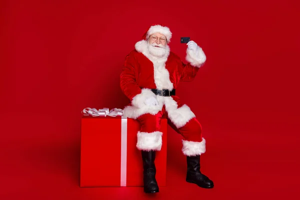 Photo of handsome positive retired man wear santa claus costume spectacles smiling buying credit card present isolated red color background — Stock Photo, Image