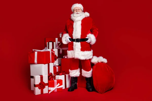 Foto van vrolijke knappe gepensioneerde man dragen kerstman kostuum bril glimlachen voorbereiding presenteert geïsoleerde rode kleur achtergrond — Stockfoto