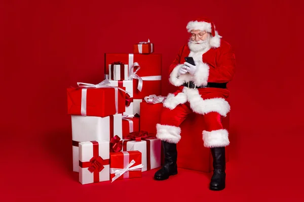 Foto de homem aposentado muito alegre usar óculos de traje de Papai Noel preparando presentes digitando dispositivo moderno isolado fundo cor vermelha — Fotografia de Stock