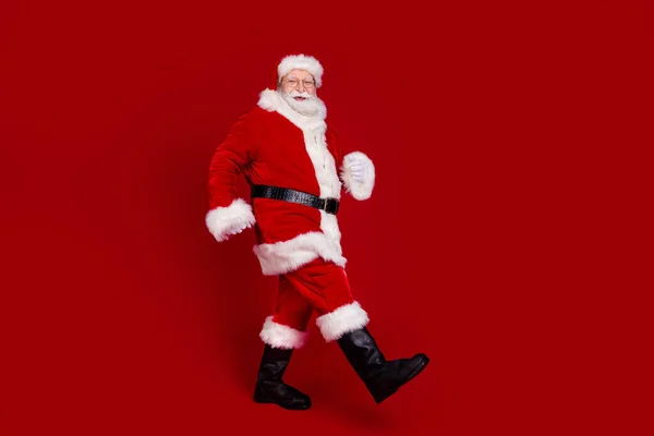 Foto de lindo hombre funky pensionista vestido de santa claus botas de traje caminando espacio vacío sonriendo aislado color rojo fondo — Foto de Stock