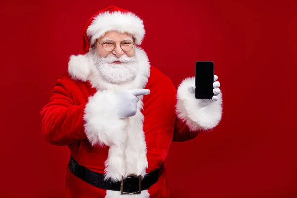 Foto de pensionista bonito homem funky vestido traje de Papai Noel apontando dispositivo moderno espaço vazio isolado fundo cor vermelha — Fotografia de Stock