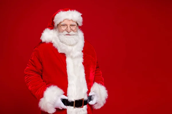 Foto de hombre jubilado alegre positivo usar traje de Santa Claus abrigo de piel cinturón gafas sonrientes brazos manos cintura aislado color rojo fondo —  Fotos de Stock