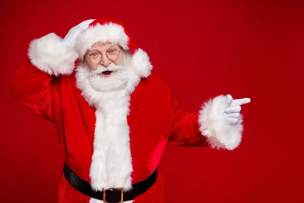 Foto di bello impressionato uomo pensionato vestito Babbo Natale costume indicando dito vuoto spazio isolato colore rosso sfondo — Foto Stock