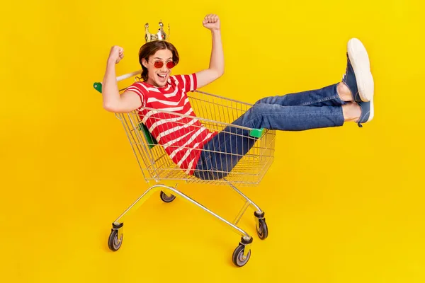 Portret van aantrekkelijke trendy vrolijke man rijden in winkelwagen gek hebben plezier geïsoleerd over helder gele kleur achtergrond — Stockfoto