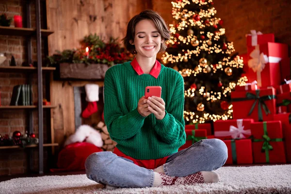 Foto de bastante brillante joven mujer vestida de punto suéter sentado piernas cruzadas chateando dispositivo moderno sonriendo en el interior habitación casa —  Fotos de Stock