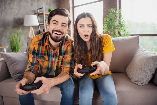 一对有趣的游戏夫妇拿着游戏机玩电子游戏享受周末穿着休闲装坐在家里沙发上的照片 — 图库照片