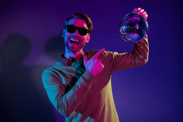 Foto retrato del hombre sonriendo en gafas de sol señalando la música disco bola dedo aislado color púrpura fondo —  Fotos de Stock