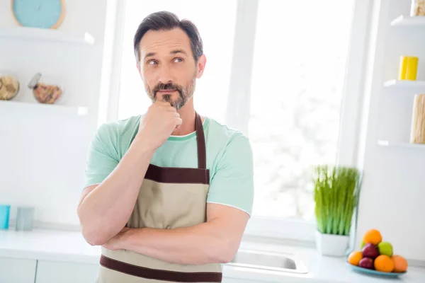 Foto de divertido hombre de edad pensativo vestido azul camiseta delantal cocina sonriendo en el interior de la habitación de casa — Foto de Stock