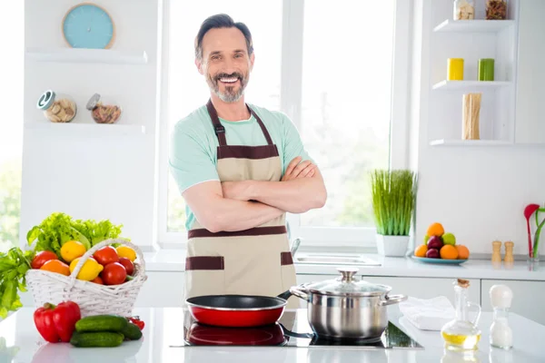 Foto de encantador hombre maduro seguro de usar delantal azul camiseta brazos de cocina cruzados sonriendo en el interior habitación de la casa — Foto de Stock