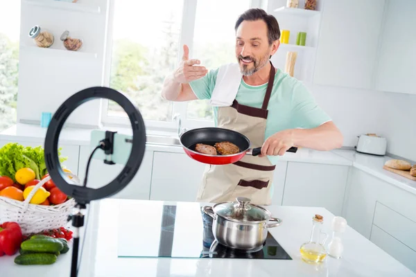 Blogcu adamın fotoğrafı telefon yayını yapıyor kızarmış et kokusu alıyor mutfak evinde mutfak önlüğü giyiyor. — Stok fotoğraf