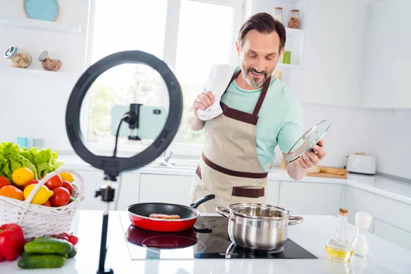 Foto de profesional divertido hombre de edad vestido azul delantal camiseta cocinar sopa hirviendo en el interior habitación de casa — Foto de Stock