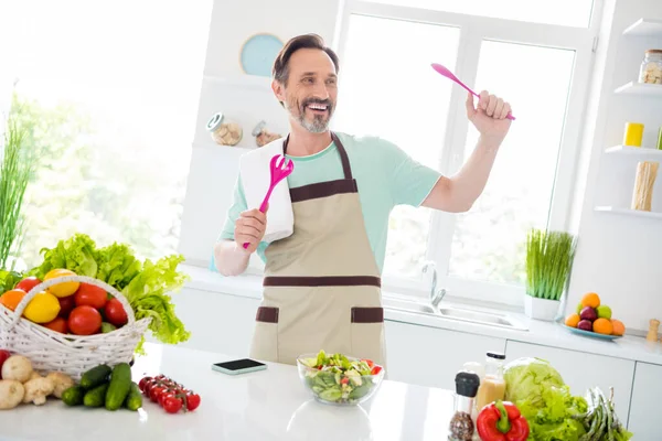 Neşeli, kaygısız, mavi tişörtlü bir adamın fotoğrafı. Gülümseyen bir önlük. Salata pişiriyor, evde dans ediyor. — Stok fotoğraf
