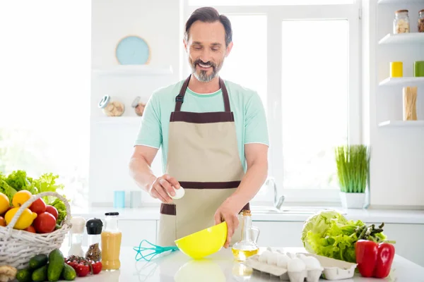 Neşeli, pozitif, olgun bir adamın fotoğrafı. Mavi tişört giyip, ev içinde omlet pişirirken gülümsüyor. — Stok fotoğraf