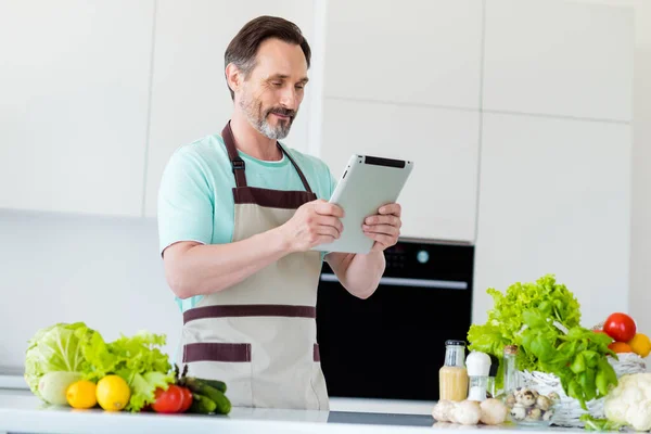 Foto de encantador hombre de edad bonita vestido azul camiseta leyendo dispositivo moderno sonriendo en el interior de la habitación de casa — Foto de Stock